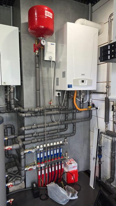 The image shows a boiler room with a Baxi brand gas boiler, a red expansion tank, and a complex network of insulated pipes. There are also several valves and gauges visible. A white cabinet is mounted on the wall to the left of the boiler. On the floor, there are some boxes, a plastic bag, and a battery. The room has gray tiled walls. The overall impression is of a functional but somewhat cluttered utility space, typical of a residential or small commercial heating system. The specific location within Кирилловское, Russia is not determinable from the visual information provided in the image itself.