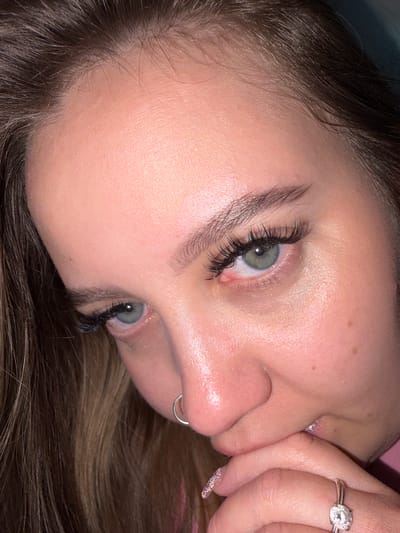 A close-up, angled view of a woman's face. She has fair skin, light brown hair, and striking green eyes enhanced with prominent eyelash extensions. A silver nose ring is visible in her left nostril. Her manicured fingers are partially obscuring her mouth, and a diamond ring is visible on her right ring finger. The background is blurred and suggests a soft, pink surface. There is no indication of Port-Katon, Russia.