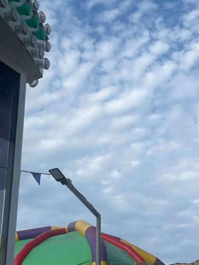The image shows a cloudy sky with some colorful inflatable structures in the foreground. A portion of a building with lights on top is visible on the left. A street lamp is also visible. The lower part of the image contains a green inflatable structure with red, yellow, and purple accents. The building on the left appears to be part of a fairground or amusement park. The overall scene suggests an outdoor recreational area.