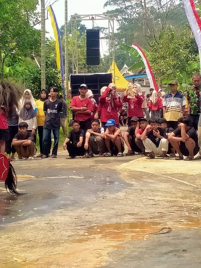 The image captures an outdoor community event in Tamansari, Indonesia, featuring approximately two dozen individuals gathered as spectators. In the immediate foreground, ten young males are squatting in a line on a wet, paved surface, facing left, observing an unseen subject. Behind them, a mixed group of standing adults, including men and women, also observes the activity. Two women wearing red shirts and hijabs are actively holding up smartphones, seemingly recording or photographing. Partially visible on the far left is a person in a performance costume, including a dark, shaggy headpiece and a red, decorated tail-like object. In the mid-ground, a temporary sound system is erected, featuring a metal truss supporting two black line array speakers, with two additional large black speakers on the ground. Two yellow canopy tents and a partial blue structure are visible in the background. Vertical banners, one yellow and blue, and another red and white (Indonesian flag colors), are attached to poles. The overall background consists of dense green trees and foliage. The paved ground exhibits multiple wet patches and scattered light-colored debris.