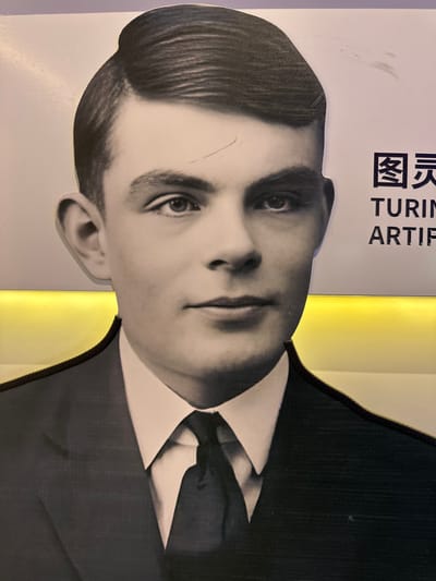 A two-dimensional, monochrome cutout of Alan Turing is prominently displayed, depicting him from the chest up. Turing wears a dark suit, a white collared shirt, and a dark tie, with his hair neatly styled and parted on his left. He presents a neutral expression, with his gaze directed slightly to the viewer's right. The cutout is bordered by a thin black outline. Behind the figure, a light-toned surface is visible, punctuated by a horizontal strip of warm yellow illumination positioned behind Turing's chest level. To the upper right of the cutout, partially visible text includes Chinese characters "图灵" (Tú Líng), followed by "TURING" and "ARTIF". Visible surface abrasions and scuff marks are present on the print, notably across Turing's forehead and in the upper right background area. This scene is located indoors, in Guangming District, China.