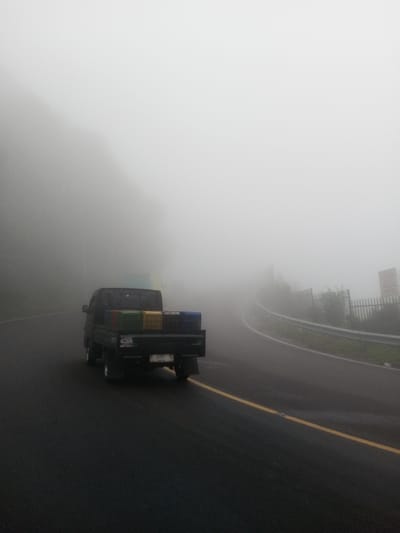 The image depicts a dark-colored pickup truck traveling on a wet asphalt road through dense fog in Siruppiddy, Sri Lanka. The truck, viewed from the rear, carries multiple rectangular crates of green, yellow, and blue colors on its flatbed. The license plate reads "B 701 YZ", and the letters "CQR" are visible on the truck's tailgate. A continuous yellow line marks the road, which curves to the right into the heavy fog. On the right side of the road, a metal guardrail is present, beyond which indistinct vegetation and a partially visible sign are observed. The left side of the road features a dark, obscured landmass, likely a hillside or dense foliage. No individuals are visible. Visibility is severely limited due to the widespread fog.