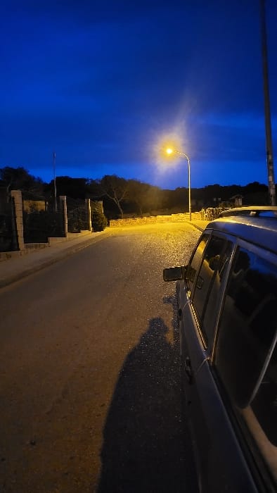 The image depicts a nighttime street scene in Santa Ponça, Spain, under a dark blue sky. A light-colored station wagon or SUV is parked parallel to a wet asphalt road on the right side of the frame. Its driver's side is visible, including the side mirror. An elongated human shadow is cast from the viewer's position onto the asphalt beside the vehicle. A single, bright yellow-orange streetlamp on a tall pole illuminates the foreground and midground, causing reflections on the damp road surface. To the left of the road, a low stone wall with concrete posts and a dark fence or railing runs parallel to the street. Behind this barrier, dense, dark vegetation, including trees and shrubs, is visible. The background consists of silhouetted hills covered in vegetation against the deep blue sky, suggesting a natural or semi-rural environment. No other individuals or explicit interactions are visible.
