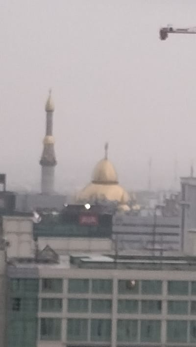 The image depicts a cityscape on a hazy day, likely in the City of Medan, Indonesia. In the background, a minaret with a gold-tipped spire stands prominently, suggesting a mosque. To its right, a large golden dome is visible, also indicative of religious architecture. The middle ground features several buildings, one with a red sign that is partially obscured. In the foreground, a modern building with a grid-like pattern of windows is shown. A construction crane is visible in the upper right corner of the image, suggesting ongoing development. The overall atmosphere is muted due to the haze.