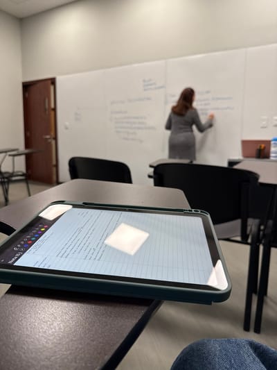 The scene depicts an indoor classroom or lecture environment located in Tizapan, Mexico. In the immediate foreground, a dark-colored tablet encased in a green protective cover rests on a dark brown desk surface, displaying digital notes with a title "Nota (5)" and prominent reflections from overhead lighting. The lower right corner reveals a portion of a person's legs, clad in blue jeans, indicating the photographer's seated position. In the midground, multiple black chairs with attached dark brown desks are arranged, predominantly unoccupied. A dark brown wooden door with a silver handle is visible on the left. In the background, a person with shoulder-length brown hair, wearing a gray dress or tunic, stands with their back to the viewer, writing on a large white board covered with blue marker text and diagrams. To the right of the whiteboard, a small, light brown container holds a blue water bottle and other objects on a desk.