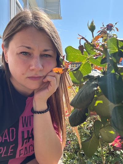 A medium shot portrait of a woman is captured outdoors in Uyutnoye, Ukraine, on a sunny day. The woman, with shoulder-length brown hair, is positioned on the left side of the frame, resting her chin on her right hand. She wears a black and pink t-shirt with white text that reads "BAD HABITS PEOPLE FACE". Her left earlobe sports a small earring. In the foreground and to the right of the woman, a rose bush with green leaves and a budding flower is visible, partially obscuring her. The background reveals a clear blue sky, the facade of a building with windows, and more of the rose garden. The lighting suggests it is daytime.