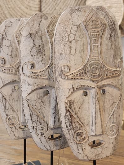 The media file displays a close-up, overhead view of three carved wooden objects stacked on top of each other. The objects are oval-shaped and feature intricate, abstract carvings with swirling patterns and geometric shapes. The wood appears weathered and has a light, distressed finish, with darker areas accentuating the carved details. Each object has two circular holes, suggesting they might be intended for hanging or for some other functional purpose. The background is softly blurred, hinting at a rustic or artisanal setting, possibly a market stall or workshop, with woven textures and wooden elements visible. The lighting suggests it is daytime, with natural light illuminating the carved surfaces and highlighting the textures of the wood. No text is visible in the image. The overall impression is one of handcrafted artistry and a natural, earthy aesthetic. The setting appears to be Maitland, United States of America.