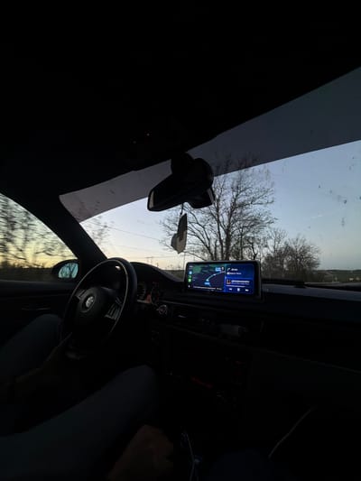 The image depicts the interior of a car driving in Boryana, Bulgaria. The driver's hands are visible on the steering wheel. A navigation device is mounted on the dashboard displaying a map and route information in Cyrillic script with "ЕВРОММИОНЕРА" prominent on the screen. Through the windshield, the view is of bare trees against a pale sky. The rearview mirror is also visible.