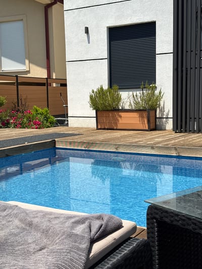 The image depicts a modern residential outdoor area with a swimming pool. The pool has bright blue tiled walls and a clear water surface reflecting the sunlight. Surrounding the pool is a wooden deck. To the left, a beige-colored building with a white-slatted window and a dark red downspout is visible. Adjacent to this building, a horizontal wooden fence runs along the property line, with green and pink flowering bushes and dark red foliage in front of it.

In the background, a white building with horizontal grey lines and a dark grey roller shutter window is prominent. A rectangular wooden planter box containing two green shrubs is placed on the deck in front of this building. To the right of the white building, a tall, dark grey vertical slatted fence or screen is situated.

In the foreground, a grey towel is draped over a lounge chair, positioned next to a dark wicker-style table with a glass top. The overall scene suggests a private, well-maintained outdoor leisure space, likely in a sunny climate. The location is identified as Lastva Grbaljska, Montenegro, implying a coastal or near-coastal setting with Mediterranean architecture and vegetation. There are no people visible in the image.