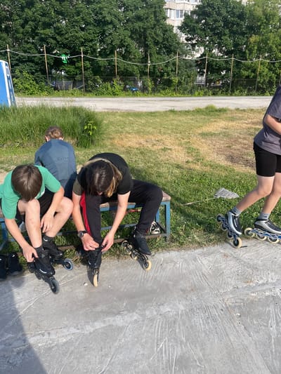 This image shows a group of three young people, two girls and one boy, sitting on a bench and the concrete ground outdoors on a sunny day. They appear to be in the process of putting on or adjusting their rollerblades.
The scene is set in an urban park or recreational area. In the background, there are tall green trees, a chain-link fence, and apartment buildings. The ground is a mix of dry, yellowed grass, green grass, and a concrete pathway. The time of day is clearly daytime, indicated by the bright sunlight casting shadows. The weather appears to be warm and sunny, typical of a summer day in Tula, Russia.
The subjects are dressed casually in t-shirts and pants. One girl is wearing black pants and a black t-shirt, and is focused on adjusting her rollerblade. Another girl is wearing black shorts and a gray t-shirt, with her legs and rollerblades visible in the upper part of the frame. The boy is wearing a green t-shirt and black pants, and is also engaged with his rollerblades. Their body language suggests concentration and perhaps a bit of casual conversation among them.
The rollerblades are a prominent object in the scene, with various models visible. One pair has a blue and white frame, and another set appears to be all black. On one of the rollerblades, the word "WAVEKON" is visible.
The overall atmosphere is relaxed and suggests a recreational activity about to begin or just finished. The presence of apartment buildings and the specific type of fence might offer clues to the precise location within Tula, Russia, but without more information, it's difficult to pinpoint.