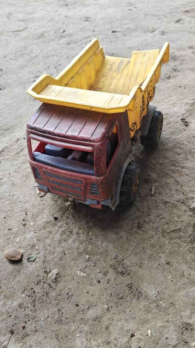 An overhead shot of a weathered, red and yellow toy dump truck sits on a dirt surface. The truck's cab is red with blue seats visible through the windows. The truck bed is yellow and appears to be stained and dirty. The tires are black and appear to be made of rubber. The ground is composed of light brown dirt with small pebbles and debris scattered across it. In the lower left corner of the image, there is a small, brown, oval-shaped object, possibly a seed or a small stone, and a single green leaf. The background is out of focus but appears to be more of the same dirt surface.