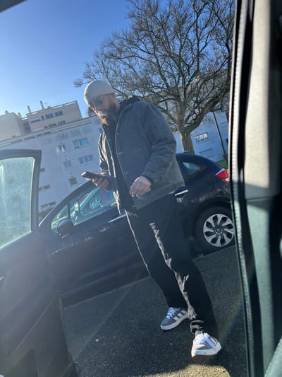 The scene depicts an outdoor urban environment in Hérouville-Saint-Clair, France, under a clear blue sky. A male individual stands in the mid-ground on asphalt pavement. He wears a light-colored beanie, sunglasses, a dark hooded jacket, black trousers, and grey and white Adidas sneakers. He has a dark beard. He holds a dark-colored smartphone in his left hand, appearing to observe its screen. A lit cigarette is held in his right hand, with smoke visibly emanating. To the left of the individual, the driver's side front door of a dark-colored car is open. Behind him, a dark-colored hatchback, consistent with a Citroën C3, is parked. In the background, a large deciduous tree with bare branches is prominent, flanked by multi-story residential buildings with light-colored facades. The scene is illuminated by bright sunlight.