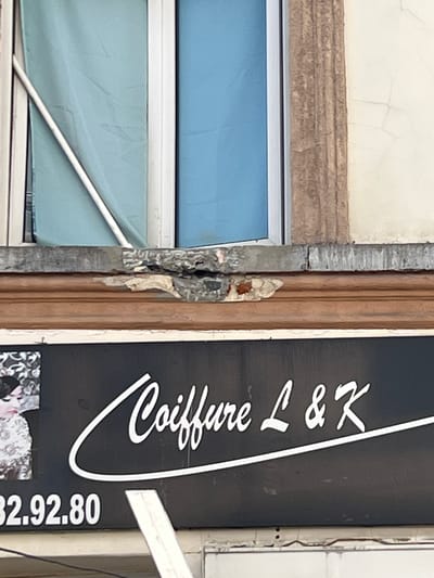 The image shows a close-up of a damaged facade above a storefront sign. A dark sign with white lettering reads "Coiffure L & K" and a phone number "32.92.80". There is also a portion of a black and white image of a person on the left side of the sign. Above the sign, there is a decorative wooden trim, and above that, the facade shows signs of significant damage, with crumbling plaster and exposed brickwork. In the background, there is a window with blue and teal colored curtains visible. The context provided indicates this scene is located in Molenbeek-Saint-Jean - Sint-Jans-Molenbeek, Belgium.