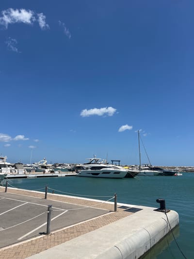 The image shows a marina in M'Diq, Morocco, on a sunny day. Several yachts of various sizes are docked alongside a concrete pier, with mooring posts and painted parking lanes. The water is calm and clear. The sky is blue with scattered white clouds. No people are visible.