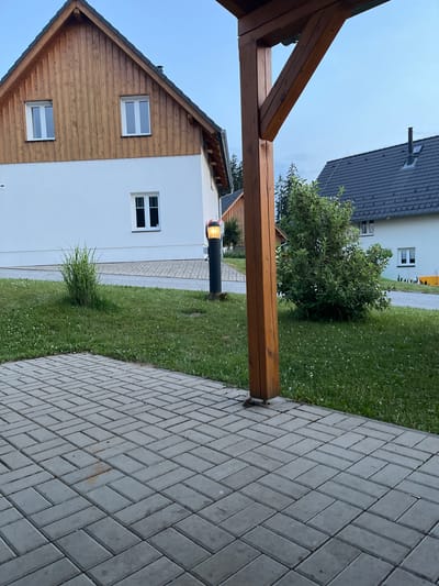 This image depicts an outdoor scene near a house in Milná, Czechia. The foreground features a wooden porch structure with visible beams and a post. To the right of the porch, a paved walkway made of gray rectangular bricks is laid in a herringbone pattern. Beyond the walkway, a lush green lawn with scattered small white flowers stretches towards a white-walled house with wooden accents. Two white-framed windows are visible on the wooden section of the house, and another on the white section. A tall, dark lamppost with a lit amber light stands near the house. In the background, another house with a dark tiled roof and a chimney can be seen, partially obscured by a large green tree. The sky is overcast and appears to be a soft, pale blue, suggesting it is either early morning or late evening, or a cloudy day. There are no people visible in the image, and the overall atmosphere is calm and serene. No text is visible in the image.