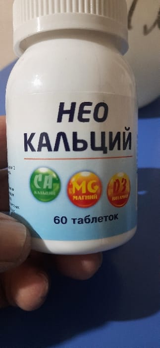 A white bottle of “NEO Calcium” supplements is in a person's hand. The label, in Cyrillic script, highlights D3 vitamin, magnesium, and calcium. It also indicates “60 tablets.” The bottle is mostly in focus, with a blurred object in the background. The backdrop is a flat, medium blue surface. The hand holding the bottle is tan.
