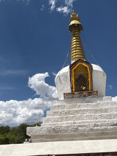 This low-angle, vertically oriented image captures a majestic white stupa, a Buddhist monument, against a brilliant blue sky interspersed with scattered white cumulus clouds. The stupa's base is composed of textured, light-grey stone blocks arranged in horizontal tiers, resembling steps. Its main body is smooth and pristine white.
Crowning the stupa is a prominent golden spire, known as a chattrāvalī, which consists of multiple stacked, disc-like elements tapering upwards to a finial. Delicate chains, possibly adorned with small bells, hang from the spire, connecting to the stupa's upper structure. Below the spire, a richly decorated rectangular section, likely the harmika, features intricate golden lattice work. This section is bordered by bright yellow garlands, possibly marigolds, and contains small clusters of colorful flowers (red, pink, and white) placed within the golden grid. To the right of this ornate panel, a vertical rectangular slab bears text in an ornate script, which appears to be Tibetan.
The bright sunlight indicates a clear daytime setting with pleasant weather. Some green foliage from trees or bushes is visible at the bottom right, suggesting a natural or landscaped environment surrounding the monument. The architectural style, particularly the golden spire and the presence of Tibetan script, strongly indicates a Tibetan Buddhist cultural site, aligning with the provided location context of Chengguan District, China. No human activity or specific event is depicted; the image focuses on the grandeur and intricate details of the sacred structure.