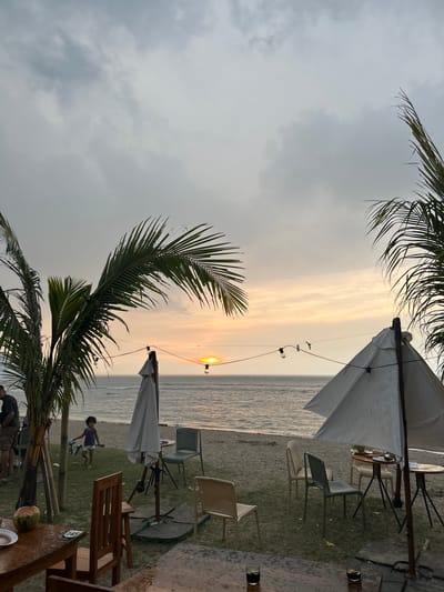 The image depicts an outdoor scene, likely a beachside restaurant or lounge in Karang Suraga, Indonesia. The time appears to be around sunset, evidenced by the sun setting over the water. Palm trees are visible framing the image on both sides, typical of a tropical beach location. There are tables and chairs arranged on a grassy area, covered with sand. Several white parasols provide shade for the seating area. Strings of lights are strung between the parasols, suggesting a relaxed, evening atmosphere. A child is riding a toy vehicle, possibly a scooter or small bike, on the grass near the tables. People are partially visible at the bottom of the frame, some standing, others sitting, near a table with a coconut drink. The ocean is in the background, with a calm surface reflecting the colors of the sunset. The sky is partially cloudy, with a gradient of colors from grey to orange near the horizon.