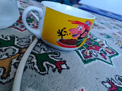 A close-up, low-angle shot shows a yellow mug with a cartoon character and a white cord. The mug is adorned with a depiction of a character with red hair and a blue hat, eating from a bowl of what appears to be cereal. The character has large blue eyes and a wide smile. The mug itself shows some signs of wear, with a chipped rim. To the left of the mug, the top portion of a white object, possibly another mug or a pitcher, is visible. A white cord runs from the bottom left of the image, curving upwards and across the scene, partially obscuring the mug. The mug and the white object rest on a patterned surface, which appears to be a rug or tablecloth. The pattern is complex, featuring geometric shapes and floral motifs in various colors including green, red, yellow, black, and white. The background is blurred, suggesting a shallow depth of field, and consists of muted colors with a hint of blue. The scene is illuminated by natural light, casting soft shadows. The location of this scene is in the city of Türkmenabat, Turkmenistan.