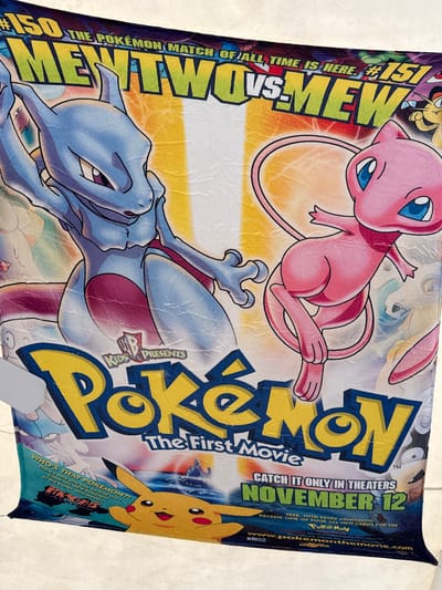 The image is a photograph of a large, creased poster, likely a movie poster for "Pokemon: The First Movie." The poster features prominent illustrations of the Pokemon characters Mewtwo and Mew, with Mewtwo on the left and Mew on the right. The title "Pokemon" is displayed in large, bold yellow letters with a blue outline. Above the character illustrations, the text "#150 MENTWO VS. MEW" is visible, indicating a significant matchup. The bottom portion of the poster displays the release date "NOVEMBER 12" in large green letters. Text related to "Kids' WB Presents" and "CATCH IT ONLY IN THEATERS" is also visible. A smaller illustration of Pikachu is present at the bottom left, alongside text describing bonus content. The poster is hung in an indoor environment, with a white wall visible in the background. The lighting suggests ambient indoor light. There is no visible indication of the city of El Paso, United States of America.