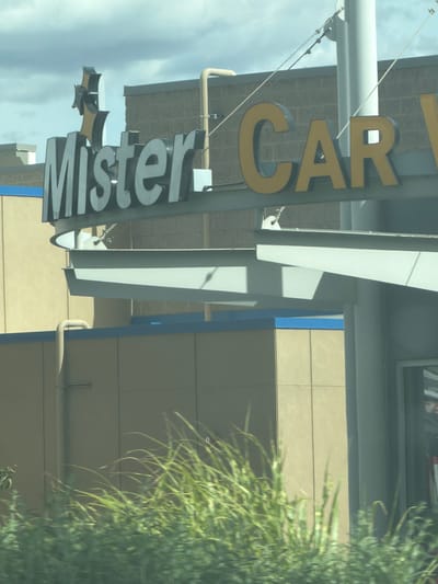 In Pasco, Washington, a "Mister Car Wash" sign is visible above a beige-colored building. The sign features large, grey, block letters spelling "Mister" on the left, and yellow, block letters spelling "CAR" on the right. A stylized, black and yellow graphic resembling a star or a propeller is positioned above the "Mister" portion of the sign. A light-colored pipe runs vertically up the side of the building behind the "Mister" sign. The building is partially obscured by a metal structure and a foreground of blurred green foliage. The sky above is cloudy.