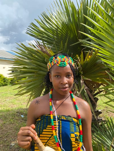 This is a portrait of a Black woman in Wang, Nigeria, outdoors during the daytime. She is wearing a colorful, traditional outfit consisting of a blue and yellow patterned dress and a beaded headband and necklace in red, green, and yellow. The beads are strung in repeating patterns. Her hair is styled in braids, and she has a tattoo of a starburst on her upper arm. She is looking towards the camera with a gentle smile.
The woman is positioned in front of lush green palm fronds, with the sky and some clouds visible in the background. A building with blue accents and some greenery can also be seen in the distance. The scene suggests a warm, possibly humid, day. The woman appears relaxed and happy. There is no visible text in the image.