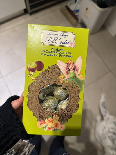 The image shows a lime green box of Marie Ange Di Costa Praline chocolates with a cream filling of pistachio. The box features an illustration of a fairy and an ornate cutout revealing the chocolates within. A hand, clad in a black knitted sweater, is holding the box. The background is cluttered with various objects, including what appears to be a white washing machine or similar appliance on the left and a patterned rug or surface in the upper right. Based on the OCR text on the box, the chocolates are an Italian product. There is no indication of the location of the image in La Lucila, Argentina.