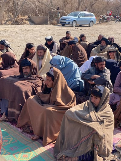 A group of men is praying outside on a rug. They are bowing in unison, dressed in traditional clothing including head coverings. A light blue car is parked nearby. The ground is barren with some trees in the background. The scene appears to be taking place in a rural or open area.
