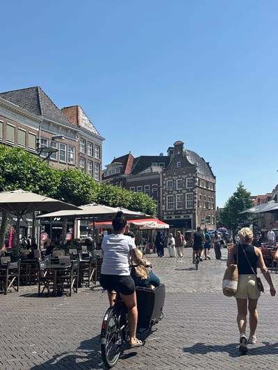 The image captures a daytime scene on a cobblestone street in the city of Zwolle, Netherlands. The street is lined with historic buildings, featuring red tile roofs and classical architectural details. Several outdoor seating areas with tables and umbrellas are set up, suggesting a commercial district with cafes or restaurants.

In the foreground, a person is riding a cargo bike, with another person seated in the front compartment of the bike. To the right, a woman with blonde hair is walking away from the camera, carrying a woven bag over her shoulder. In the background, several other people are visible walking and cycling. The sky is clear and blue, indicating sunny weather. The overall impression is that of a bustling urban environment on a warm day.