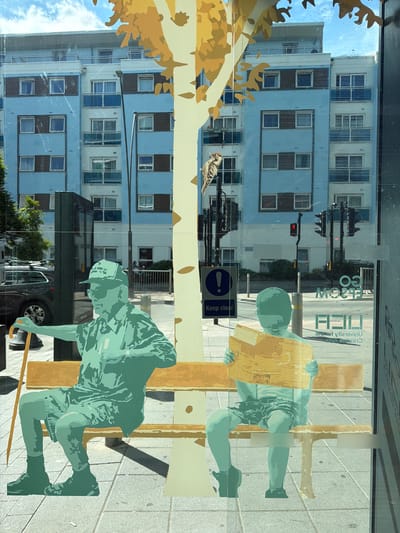 The image shows a street scene viewed through a glass window, likely in Epsom and Ewell, United Kingdom, as suggested by signage visible on the glass. The foreground features a stylized graphic art installation depicting two figures on a bench. On the left is a figure of an older person wearing a cap, sunglasses, and casual clothing, holding a walking stick. On the right is a younger figure, appearing to be a child, reading a book or newspaper. Behind the figures, a tall, stylized tree trunk with yellow leaves is visible, also part of the art.

Through the window, a modern, light blue apartment building with brown accents is prominent. The building has multiple stories, each with numerous windows and balconies with blue railings. A street with traffic lights, lampposts, and parked cars is visible in the middle ground. One traffic light shows a red signal. A sign with an exclamation mark and the text "Keep clear" is also visible on a post. Reflections on the glass obscure some details of the background, including a black car and a fence. Text, partially obscured and reversed, indicating "UNIVERSITY FOR THE CREATIVE ARTS" and "EPSOM" is visible on the glass in the lower right portion of the image. The pavement in front of the bench is paved with grey tiles. The sky above is blue with scattered white clouds.