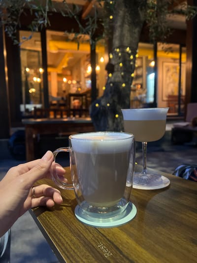 A hand with a gold ring is holding a clear mug of latte, with a foam top, on a wooden table. Next to the mug is a stemmed glass with a light-colored drink and a white foam top. In the background, a tree is decorated with fairy lights, and the blurred lights of a building are visible. The scene appears to be outdoors, possibly a cafe or restaurant, in Bjeliši, Montenegro.