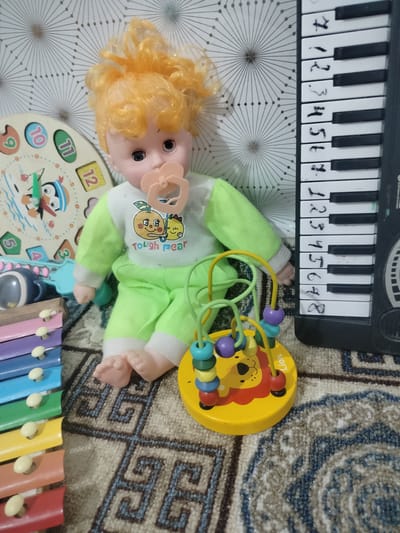 The image shows a collection of children's toys arranged on a patterned carpet. In the center is a doll with orange hair, wearing a light green and white outfit with cartoon characters on it. The doll has a pacifier in its mouth. To the left of the doll is a wooden clock toy with numbers and cartoon penguins. Next to the clock is a xylophone with colorful bars and white mallets. To the right of the doll is a black toy piano keyboard with handwritten numbers on the keys. In front of the doll and to the right is a yellow, round bead maze toy with a lion illustration. The background features a wall with a repeating geometric pattern. The overall scene depicts a child's play area, likely indoors. The provided context does not specify the location within the city of Cholpon, Kyrgyzstan.