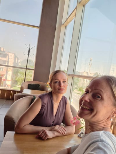 Two women are sitting at a table in what appears to be a cafe or restaurant with large windows offering a view of the city of Arseniev, Russia. The woman in the foreground is closer to the camera and is smiling broadly, looking towards the lens. She has blonde hair and is wearing a white t-shirt and pearl earrings. The woman in the background is looking towards the camera with a neutral expression. She has blonde hair and is wearing a mauve top and a delicate necklace. The table between them is made of wood, and a pink flower is visible in a vase. Outside the window, buildings, trees, and a cell tower can be seen. A coat rack with multiple hooks is positioned in front of the window.