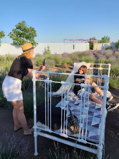 In an outdoor lavender field in Nechaiane, Ukraine, a woman wearing a black t-shirt, white shorts, and a straw hat is taking a picture of a younger person with her phone. The younger person, who appears to be a teenager, is sitting on a white metal bed with a quilted blanket. They are wearing a black t-shirt and shorts, with black sneakers. The scene is set during what appears to be late afternoon or early evening, with warm, golden sunlight casting shadows. The sky is clear blue. In the background, a white fence runs along the edge of the property, with some trees and a structure with pink decorations visible over the fence. The lavender bushes are in full bloom, creating a vibrant purple landscape. The atmosphere seems relaxed and pleasant, suggesting a photoshoot or a casual outing. The younger person is looking towards the camera with a slight smile. A leopard-print bag is placed on the bed next to them. Visible text on the sneakers reads "FILA".