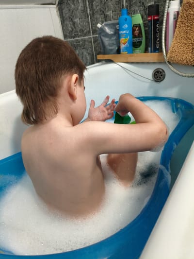 The image shows a young child, viewed from behind, sitting in a blue bathtub filled with white bubbles. The child has short, brown hair and is facing away from the camera, looking towards the wall where several bottles of shampoo and other bathroom products are placed on a wooden shelf. Among the visible products are a blue bottle with "NAMTU" written on it, a green bottle with a picture of a woman's face and "Наума" written on it, and a black and red can with "EDISON" written on it. The bathroom appears to have tiled walls. The overall scene suggests a bath time activity, likely taking place indoors, possibly in a residential bathroom in Kotovsk, Russia, as indicated by the location context. The child's arms are raised, and they appear to be holding a green object, possibly a bath toy or a bottle of soap, in their right hand. The presence of soap suds and the bath setting imply a moment of hygiene and play.