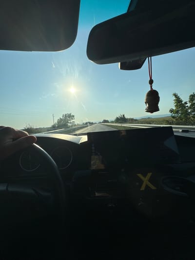The image presents a first-person perspective from the interior of a vehicle, traveling on a road in the vicinity of Mogila, Bulgaria, under a clear sky. The sun is low in the upper-left quadrant of the frame, shining directly through the windshield and causing significant lens flare.
Within the vehicle's interior, a gray sun visor is partially visible in the upper-left corner. A dark rearview mirror assembly, encompassing sensor housing, is centrally positioned at the top of the frame. A small, brown, carved ornament, resembling a Buddha head, hangs from the rearview mirror by a red cord.
A light-skinned left hand is visible, gripping the black steering wheel on the left side of the frame. Behind the steering wheel, parts of the instrument cluster are discernible, showing numerical increments reaching at least 260. The dashboard features a prominent, dark central display screen. Below this screen, a distinct yellow letter "X" is affixed to the dashboard, with a small digital display showing ".69" to its left.
The exterior environment depicts an asphalt road extending into the distance. A metal guardrail lines the right side of the road, beyond which green vegetation and fields are visible. Scattered trees and bushes populate the mid-ground, and a utility pole stands on the left side of the road. Distant, hazy hills or mountains form the horizon line under a blue sky, which lightens towards the sun.