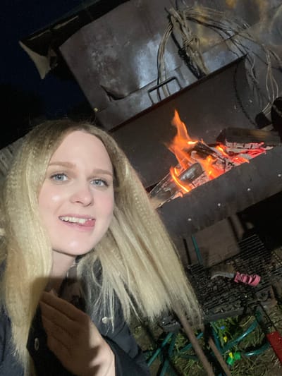 A young woman captures a selfie during what appears to be a nighttime outdoor gathering in Kauchuk Stantsiya, Russia. The image is taken from a tilted perspective, giving it a candid feel.

In the foreground, the woman smiles broadly at the camera, her blue eyes clearly visible. She has long, blonde, crimped hair that cascades across a significant portion of the lower frame. She is wearing a dark jacket, and her happy expression suggests a relaxed and enjoyable atmosphere.

To the left and slightly behind the woman, a robust fire burns brightly in a dark metal fire pit, serving as a primary light source. Orange and yellow flames engulf visible logs within the pit. Adjacent to the fire pit on the left, a dark surface with tangled wires or ropes can be seen, hinting at a rustic outdoor setup.

The background to the right of the woman reveals various outdoor objects. These include a dark metal grill, a pink-handled tool or knife hanging on the grill, and green metal structures that could be parts of outdoor furniture or equipment. Patches of green grass and dirt are visible on the ground, along with small colorful pieces of debris. The scene evokes a casual outdoor event like a campfire or barbecue. No visible text is present in the image.