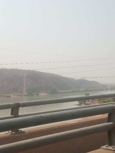 The image depicts a hazy, wide view of the Niger River from the Jebba Bridge in Jebba, Nigeria. In the immediate foreground, a grey metal bridge railing with multiple horizontal bars and vertical supports is visible, set against a concrete barrier. The river's surface, a calm greenish-brown, occupies the midground, with faint dark specks, possibly small boats or people, visible on the water. Beyond the river, on the far bank, a large, rocky hill with sparse, dry vegetation rises prominently, featuring a faint lighter-colored path or track ascending its slope. Thin, dark horizontal lines, consistent with power lines, traverse the upper midground. The sky is uniformly light grey, indicating atmospheric haze or an overcast condition. No clear individuals or specific actions are discernible.