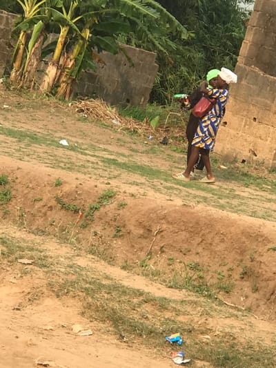 Two people are outdoors near greenery and a brick structure. One person carries the other on their back. The person being carried wears a blue patterned dress, white head covering, and carries a red purse. Both wear sandals. The ground is dirt with scattered grass. Trash, including a blue and white bag, litters the area. Banana trees grow nearby.