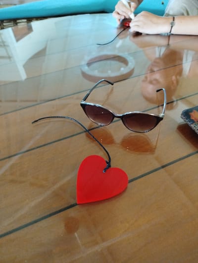 An overhead shot shows a glass table with two pairs of sunglasses, one with a red heart-shaped charm attached by a string. A person's hands are visible in the upper right corner of the image, holding a white pen and appearing to write on a red object, likely the heart charm. The table has a polished wooden surface with dark lines running across it, possibly indicating planks. In the background, the edge of a turquoise surface, possibly a pool or a countertop, is visible. The scene is set in Gulhi Falhu, Maldives, suggesting a tropical or resort environment.
