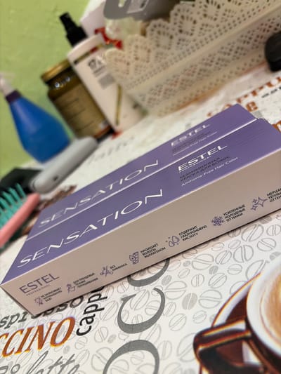 The image shows two boxes of Estel Professional Sensation hair color, which is an ammonia-free hair color. The boxes are purple and white, with the brand name and product name printed in silver and white lettering. The boxes are placed on a table with a pattern of coffee beans. In the background, there are various cosmetic products, including a blue spray bottle, a glass jar with a golden lid, and a white bottle with a black cap. The overall scene appears to be a personal collection or display of hair and beauty products, possibly in a home or salon setting. There is no indication of the specific location within the city of Kimry, Russia.