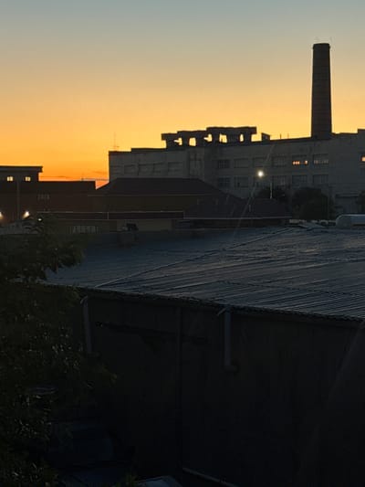 The image depicts an industrial scene at dusk, likely in Zhanaozen, Kazakhstan. The setting is dominated by large, multi-story buildings with a prominent smokestack or chimney on the left side. The architecture appears to be that of an older industrial complex, with visible windows, some of which are illuminated, suggesting occupancy or a power source.

The sky transitions from a pale blue at the top to a vibrant orange and yellow gradient towards the horizon, indicating either a sunset or sunrise. The light suggests it's either early morning or late evening.

A large, dark, corrugated roof structure runs diagonally across the right side of the image, casting a shadow over the foreground. Below this structure, there is dense foliage, possibly trees or bushes, adding a natural element to the otherwise industrial landscape. Some lights, possibly streetlights or building lights, are visible in the distance, illuminating parts of the buildings and casting faint glows.

The overall mood is somewhat somber and industrial, with the fading light creating long shadows and highlighting the starkness of the buildings. There is no visible text in the image. The scene conveys a sense of a working city or an abandoned industrial area as day transitions into night.