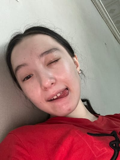 A close-up, chest-up, selfie-style photograph features a young East Asian woman with dark hair pulled back, wearing a red t-shirt. She is lying down, with her head tilted to the left and resting against a textured, beige surface. Her left eye is winking, and her right eye is open, looking towards the camera. Her mouth is slightly open, with her tongue sticking out and touching her lower lip. A small pearl earring is visible on her right ear. The background consists of a light-colored wall with a decorative molding at the top. The lighting is diffused and appears to be from an overhead source. The image is taken from a high angle, looking down at the subject. The location within Nookat, Kyrgyzstan, is not discernible from the visual information present in the image.