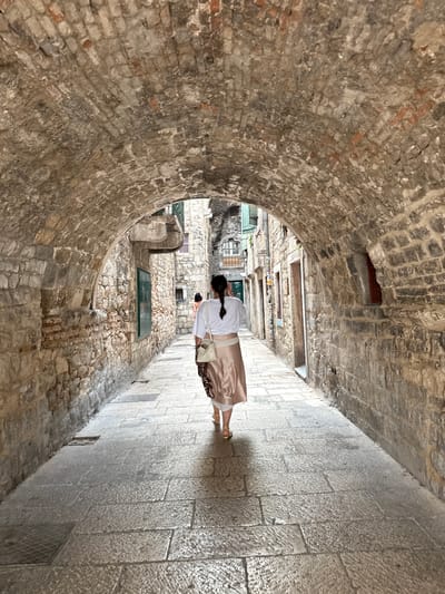 The image shows a woman from the back walking down a narrow, stone-paved alleyway in Split, Croatia. She is wearing a white top and a flowing, satin skirt. She has her hair in a braid and is carrying a light-colored purse on her shoulder. The alley is characterized by ancient stone walls on one side, with a rounded archway leading further into the passage. The other side of the alley is lined with stone buildings, and the ground is paved with large, light-colored stones. Some windows and doors are visible on the buildings. A green glass-covered opening is set into the stonework on the right side of the alley. The lighting suggests it is daytime, possibly late morning or early afternoon, with sunlight casting shadows on the ground and walls. The atmosphere appears tranquil.