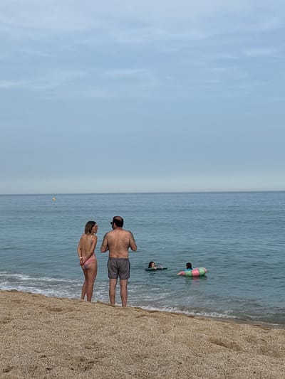 A wide shot captures a beach scene in Vilassar de Mar, Spain, under a hazy, overcast sky. The foreground consists of a pebble beach with gentle waves lapping at the shore. In the mid-ground, a couple stands near the water's edge, facing away from the camera and looking out at the sea. The woman is wearing a pink bikini, and the man is wearing dark swim shorts. Further out in the water, two children are visible. One child is floating on a green and pink inflatable ring, and another is on a small dark float or boogie board. A yellow buoy floats in the distance. The sea is a calm, muted blue-green, and the horizon is a pale line where the sea meets the sky. The overall impression is of a quiet, somewhat subdued beach day.