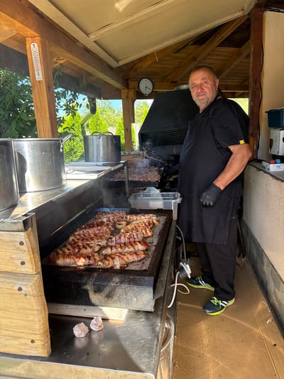 A man in an apron and black pants is standing in front of a grill where food is cooking. The grill is under a wooden shelter with a slanted roof, and the outdoor environment with trees and foliage is visible in the background. A thermometer is attached to a wooden pillar on the left side of the shelter. The man is wearing black gloves and black athletic shoes with green laces. He is looking towards the camera with a neutral expression. The cooking area appears to be a professional outdoor kitchen or food stall. The presence of a thermometer suggests it's a warm day. The location is identified as Lozno, Bulgaria, likely a public space or establishment.