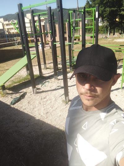A medium eye-level shot of a man in a black cap and a gray patterned t-shirt facing the camera, likely taking a selfie. He appears to be in an outdoor park or playground, evidenced by the playground equipment visible in the background, including climbing bars, rings, and a slide. The ground is covered in gravel. The park area is situated within the city of Sessa Aurunca, Italy, indicated by the buildings and landscape in the background. The scene is well-lit, suggesting a sunny day. The man is the primary subject of the photo, with the park and city as the backdrop.
