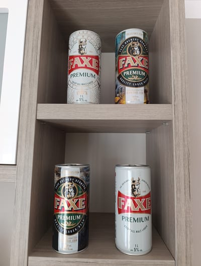 Four cans of Faxe Premium beer are displayed on two shelves of a light wood-effect cabinet. The top shelf holds two cans: one predominantly white with red and gold lettering, and another with a more colorful design featuring a viking head and a seascape. The bottom shelf also has two cans: one with a dark, patterned design featuring a viking and a seascape, and another white can similar to the one on the top shelf. The background is a plain, light-colored wall. There is no indication of the city of Toruń, Poland in the image.