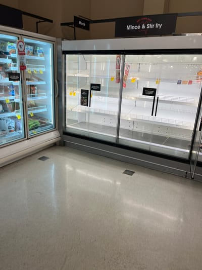 The image shows two empty refrigerated display cases in a supermarket. The top case has a sign above it that says "Mince & Stir fry". The bottom case has a sign that says "$7" and has some frozen food items inside. The floor is light-colored tile. The location of the image is a supermarket in Middle Park, Australia (latitude: -27.556, longitude: 152.923).  The image was taken at an angle, looking down at the cases. The cases are illuminated from within. The lighting in the room is bright. There is no one in the image.