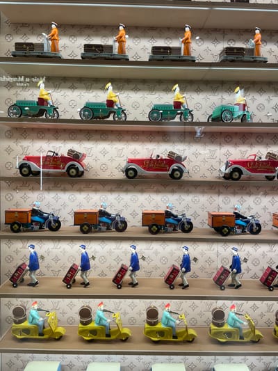 The image shows a display case with four rows of different miniature vehicles in a store. The vehicles include: a train with an attendant, a vintage green car with a driver, a vintage red car with "Louis Vuitton" written on the side, a moped with a Louis Vuitton trunk and a figurine wearing a sailor outfit, a figurine in a blue uniform pulling a rolling suitcase, and a golf cart with a driver. The vehicles appear to be merchandise. The backdrop of the display case has a Louis Vuitton logo pattern. The photo was taken at the Chengdu IFS Mall in Jinjiang District, China.